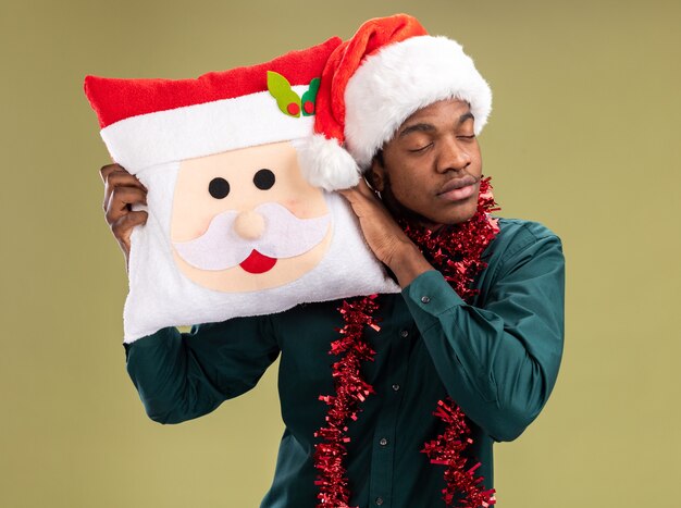 African american man in santa hat with garland holding christmas pillow with closed eyes standing over green background