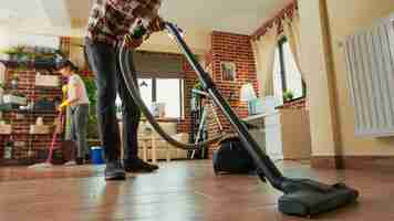 Free photo african american man vacuuming floors in living room, cleaning apartment with girlfriend. young adult using vacuum cleaner and woman wiping shelves with all purpose cleaner. tripod shot.