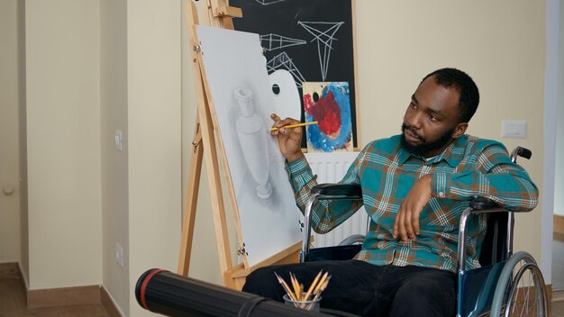 African american man with disability using pencil to draw on canvas, learning new artistic skills in art class with teacher. Young student in wheelchair drawing skethc masterpiece in workshop.