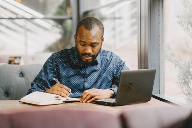 African-american man working behind a laptop and writing in a notebook. Man with beard sitting in a cafe.