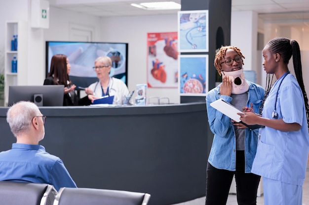 Free photo african american nurse and patient doing checkup visit in waiting room lobby, wearing cervical neck collar after accident injury. medical assistant consulting injured woman at healthcare facility.