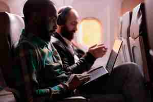 Free photo african american passenger working on laptop during sunset flight, waiting to arrive at holiday destination. flying in economy class with group of tourists, using computer online.