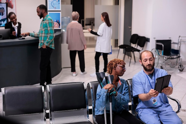 Free photo african american patient and nurse looking at tablet to find dsease diagnosis on digital device, suffering from physical impairment in waiting area. woman and specialist in hospital lobby.