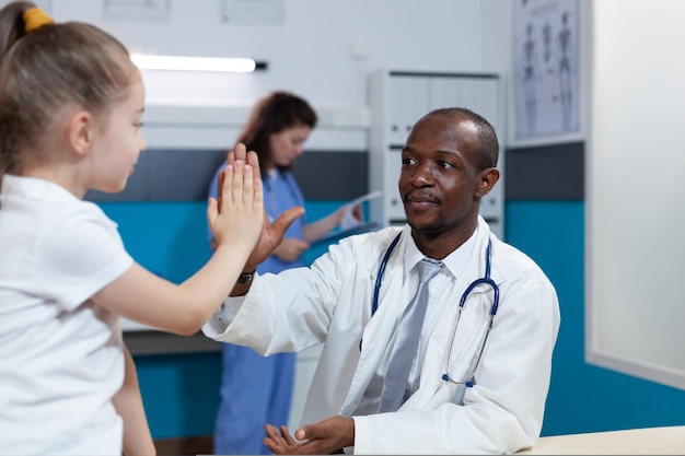 African american pediatrician doctor giving high five to young children during medical appointment in hospital office. Therapist man explaining illness symptoms discussing healthcare treatment