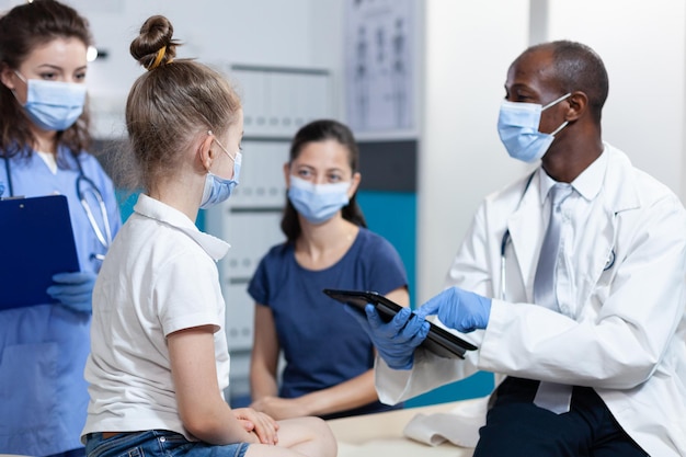 African american pediatrician doctor with protection face mask against coronavirus explaining medical treatment to family during clinical appointment in hospital office. Health care service