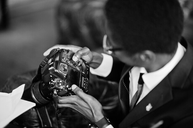 African american photographer paparazzi man wear on black suit and glasses sitting at office with camera and working behind laptop