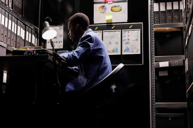 Free photo african american police officer working at crime case late at night, sitting at desk in police station. private detective looking at criminal investigations files, analyzing suspect information