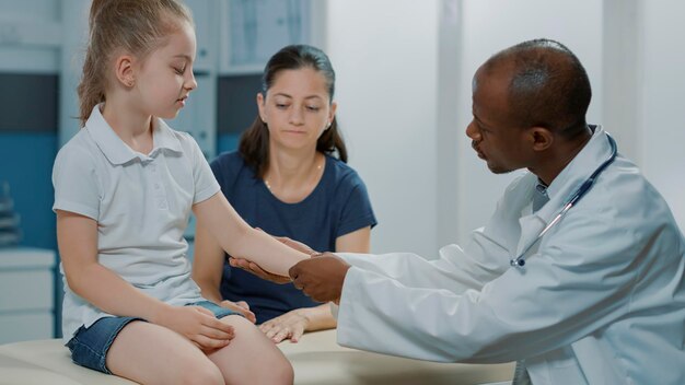 African american specialist examining injury of little kid in cabinet. General practitioner doing consultation on small girl with fractured arm in pain, giving assistance and medical support.