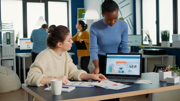 African american woman holding laptop asking colleague looking at clipboard with charts for advice regarding financial data in busy office. Two colleagues analzying market share brainstorming ideas.