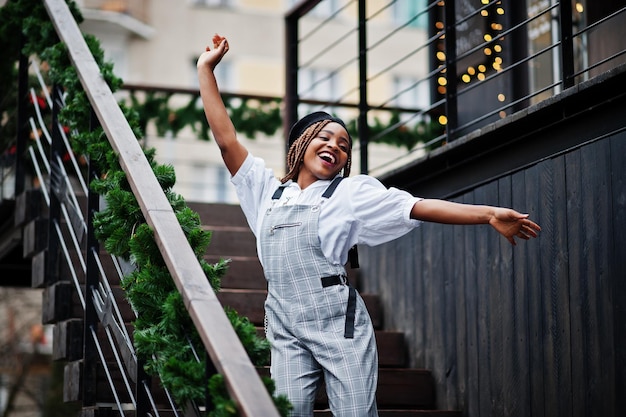 African american woman in overalls and beret posed in outdoor terrace with christmas decorations garland