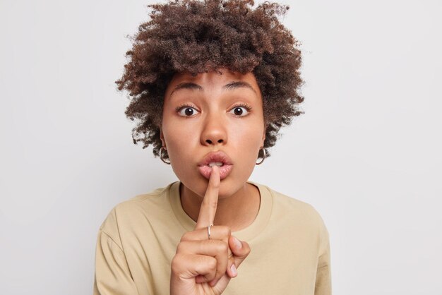 African American woman shows hush sign has mysterious expression asks to zip your lips tells you secret information dressed in beige t shirt isolated over white background. Keep silence please