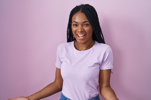 African american woman with braids standing over pink background smiling cheerful with open arms as friendly welcome, positive and confident greetings