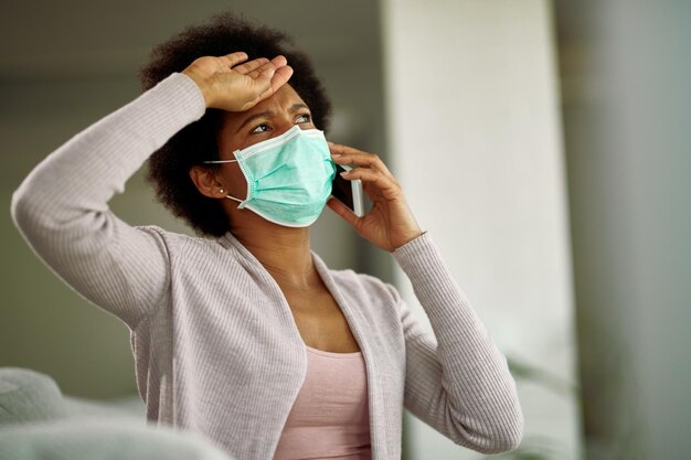 African American woman with face mask feeling worried while talking on cell phone at home