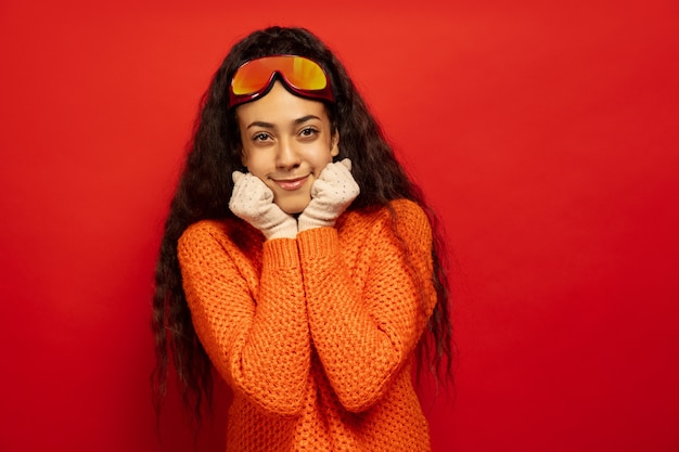 Free photo african-american young brunette woman's portrait in ski mask on red studio background. concept of human emotions, facial expression, sales, ad, winter sport and holidays. smiling, looks cute.