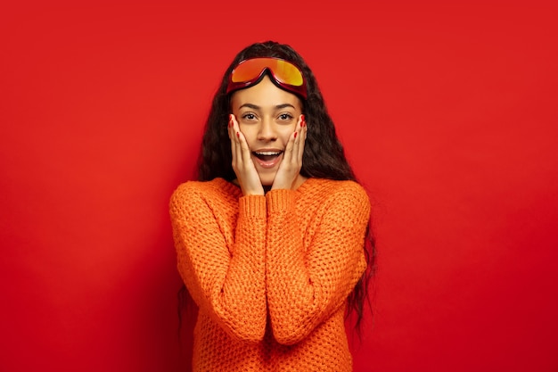 Free photo african-american young brunette woman's portrait in ski mask on red studio background. concept of human emotions, facial expression, sales, ad, winter sport and holidays. surprised, astonished.