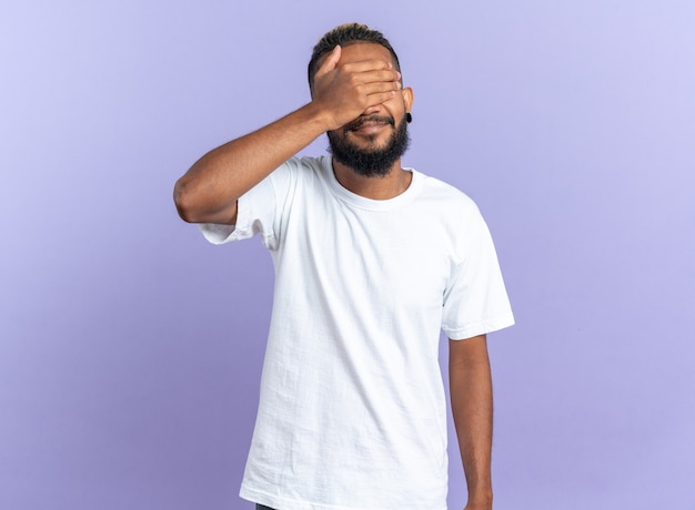 African american young man in white t-shirt closing eyes with hand standing over blue background