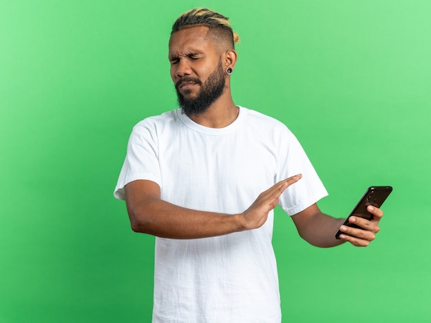 African american young man in white t-shirt holding smartphone making defense gesture with disgusted expression worried standing over green background