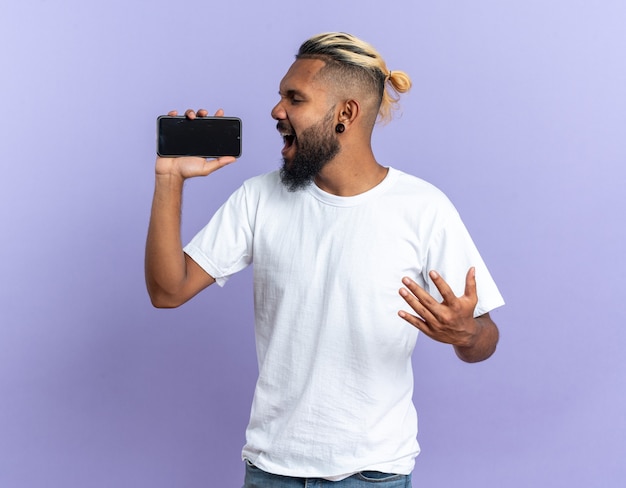 African american young man in white t-shirt holding smartphone using as microphone singing happy and emotional