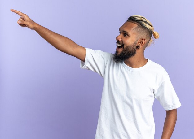 African american young man in white t-shirt looking aside happy and cheerful pointing with index finger at something standing over blue background