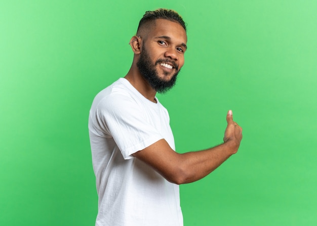 African american young man in white t-shirt looking at camera smiling cheerfully pointing back with finger standing over green background