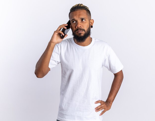 African american young man in white t-shirt looking confused while talking on mobile phone 