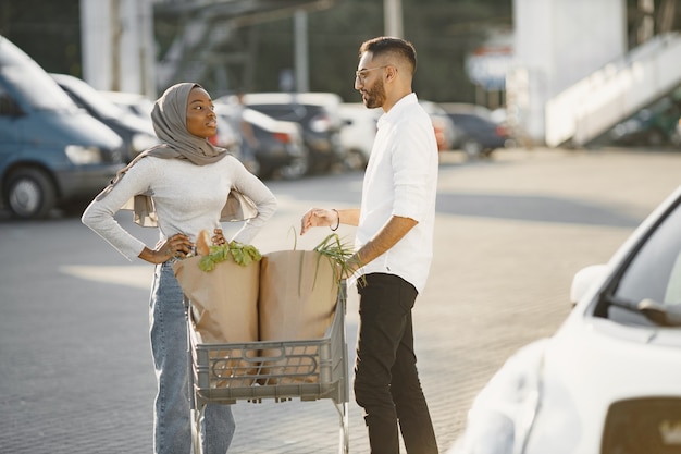 African Arabian couple stands with groceries near electric car. Charging electric car at the electric gas station