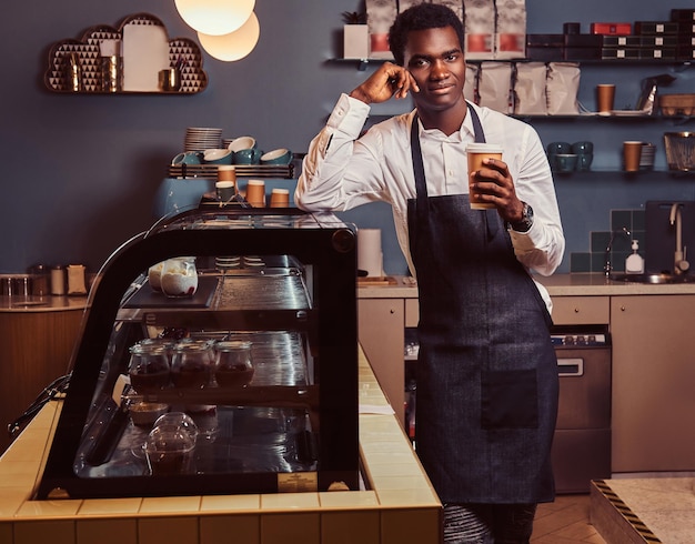 Free photo african barista smiling at camera relaxing after workday with coffee while leaning on counter at coffee shop.