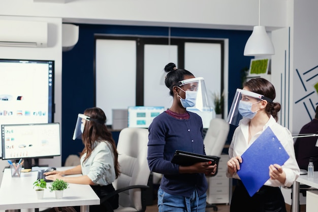 Free Photo african and caucasian coworkers discussing statistics in workplace wearing face mask. multiethnic business team working respecting social distance during global pandemic with coronavirus.