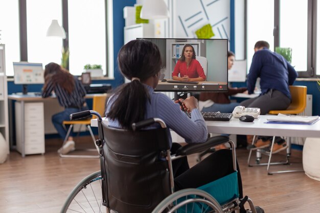 African disabled handicapped businesswoman sitting immobilized in wheelchair talking with remote partner on video call from startup business office