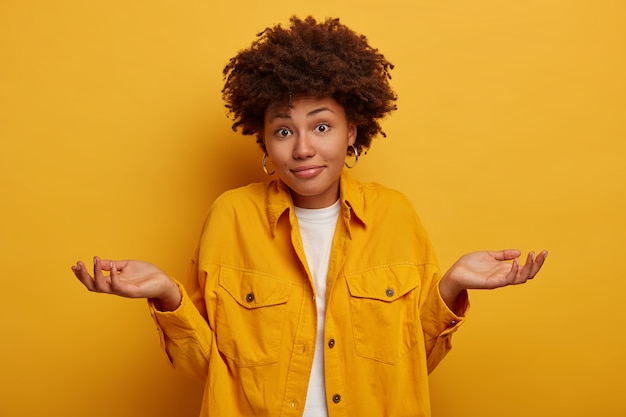 African lady shrugs shoulders and expresses uncertainty, makes decision, wears fashionable clothing, spreads palms sideways isolated on yellow wall.