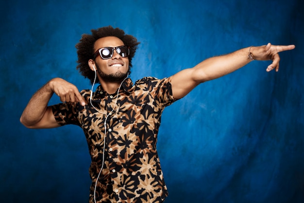 Free Photo african man listening music in headphones, dancing over blue wall.