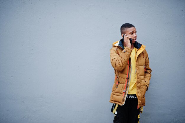 African man wear on jacket at cold weather posed outdoor against grey wall speaking on mobile phone