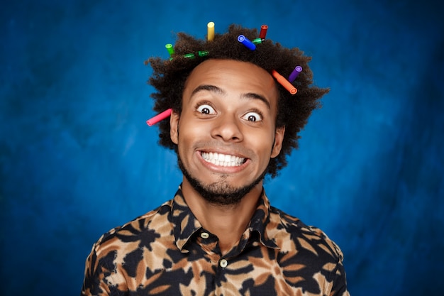 African man with markers in hair smiling over blue wall.