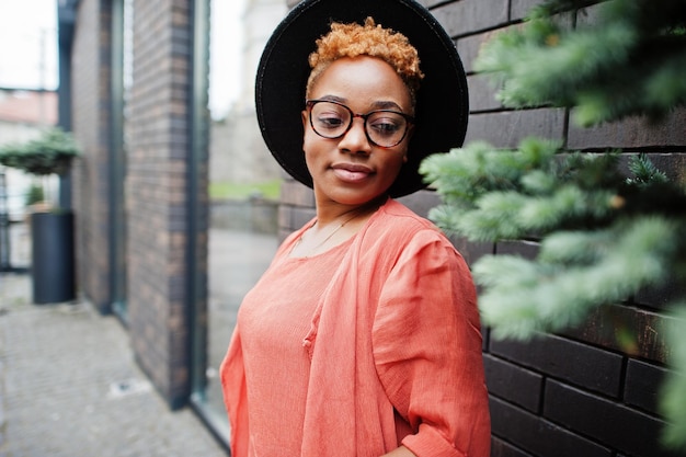 Free photo african red haired woman wear eyeglasses orange dress and black hat posing outdoor