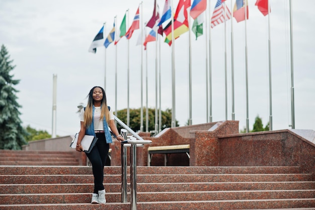 African student female posed with backpack and school items on yard of university against flags of different countries