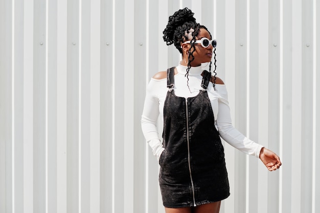 African woman in denim overalls skirt white sunglasses posed against white steel wall