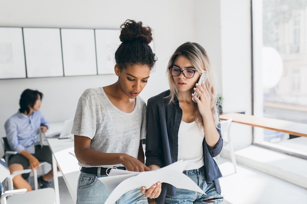 African woman in white t-shirt found mistake in the report. Worried female office worker calling colleague to solve working problems while her co-workers doing their job.