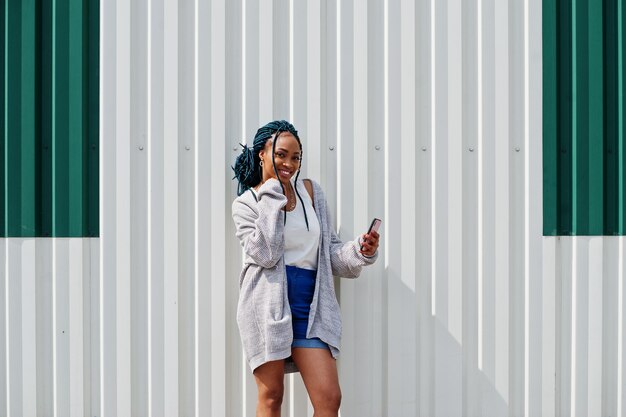 African woman with dreads hair in jeans shorts posed against white steel wall with mobile phone in hand