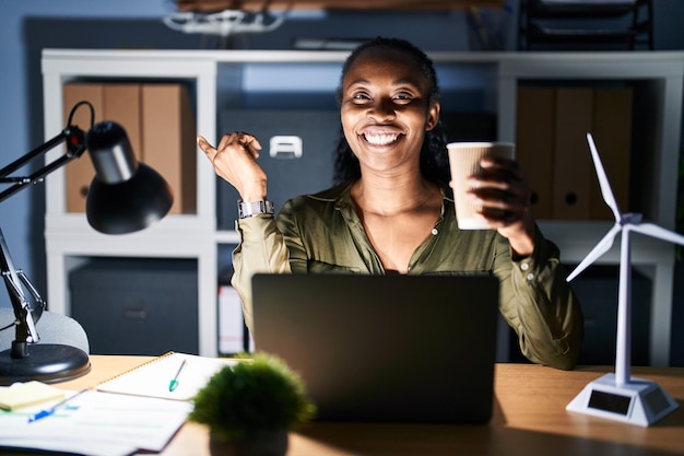 Free Photo african woman working using computer laptop at night with a big smile on face pointing with hand finger to the side looking at the camera