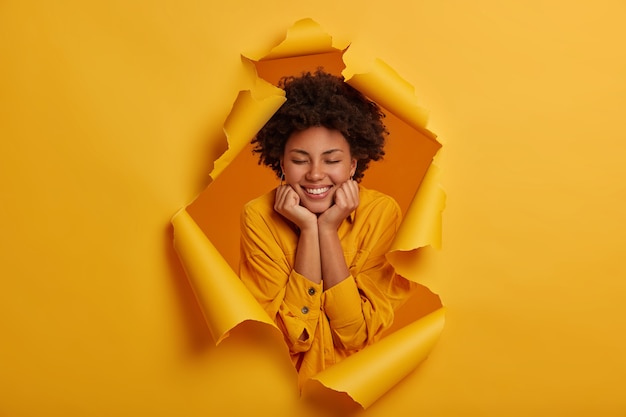 Afro American joyful woman enjoys pleasant communication, smiles broadly, holds both hands under chin, stands with closed eyes