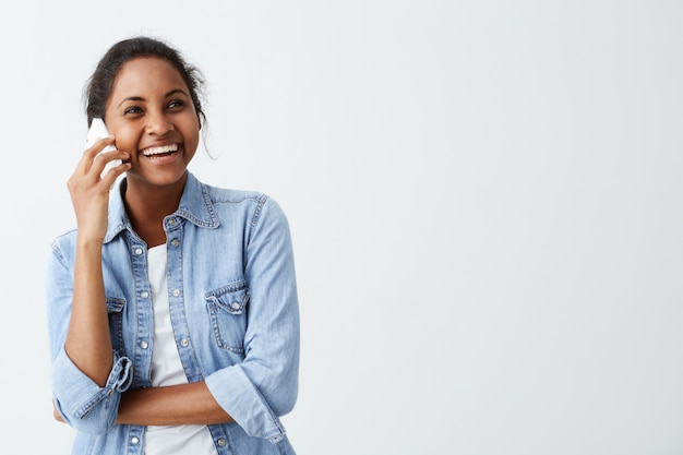 Afro-american young glad woman dressed in blue shirt over white t-shirt having conversation over smart phone, laughing, sharing good news with her friends. People and positive emotions.