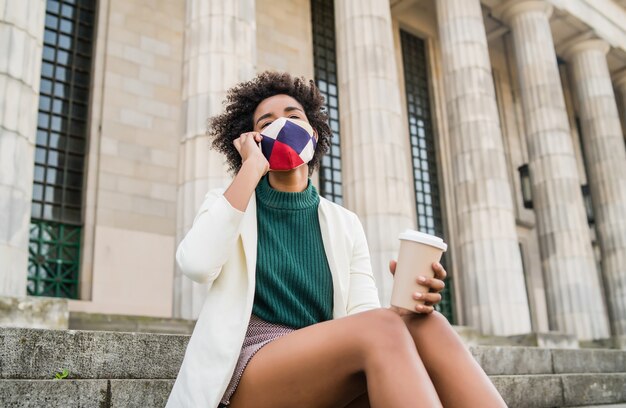 Afro business woman wearing protective mask and talking on the phone while sitting on stairs outdoors at the street. Business and urban concept.