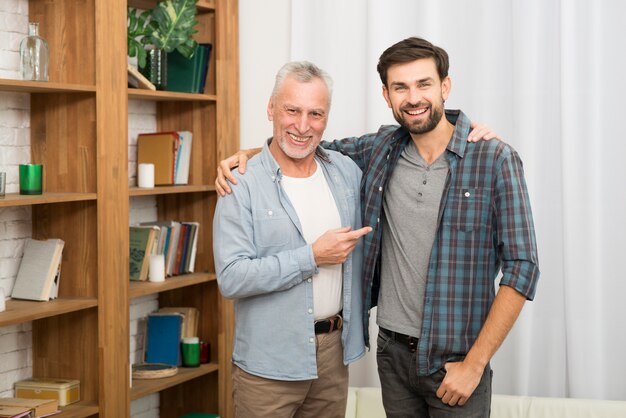 Aged happy man pointing and hugging with young smiling guy in room