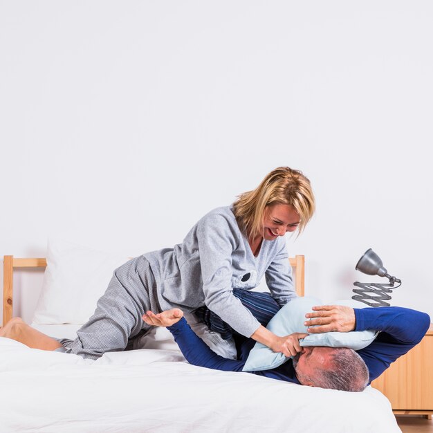 Aged happy woman and man having fun with pillows on bed in bedroom