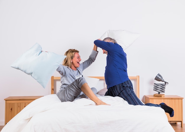 Free photo aged happy woman and man with pillows having fun on bed in bedroom