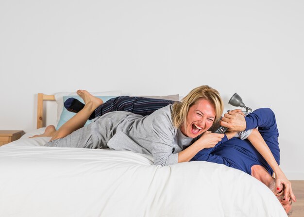 Aged laughing woman wanting to take TV remote from man hands on bed