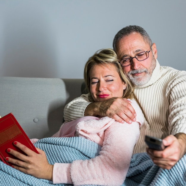 Aged man with TV remote watching TV and cheerful woman with book on sofa