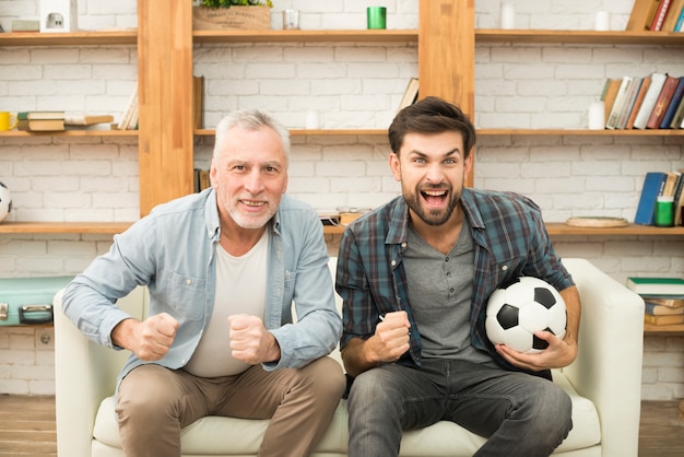 Aged man and young crying guy with ball watching TV on sofa