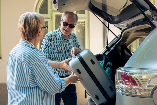 Aged people loading trolley in car trunk, travelling on retirement holiday vacation during summer. Senior people leaving on road trip adventure, putting suitcase and travel bags.