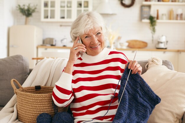 Aged people, retirement, leisure and modern technology concept. Beautiful happy grandma with gray hair talking to her granddaughter on mobile phone while knitting scarf on couch in living room